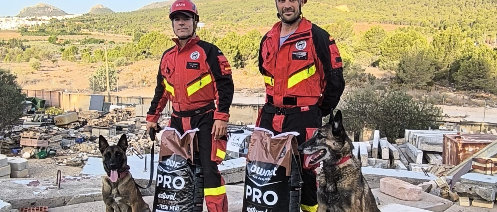 Dos guías caninos posando con sus perros. Delante de ellos y a sus pies hay dos sacos de pienso de la marca OWNAT.