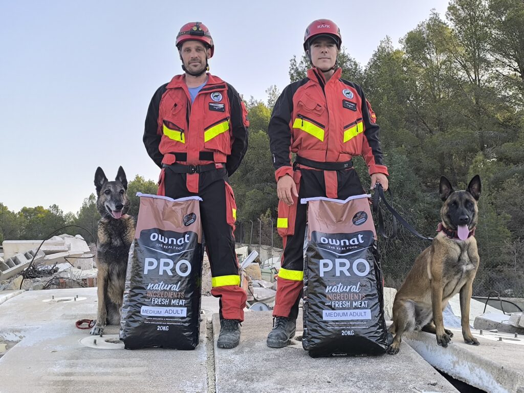 Entreno Unidad Canina K9.
Dos guías caninos junto a sus perros de búsqueda. A los pies de los guías hay dos sacos de pienso para perros marca OWNAT PRO